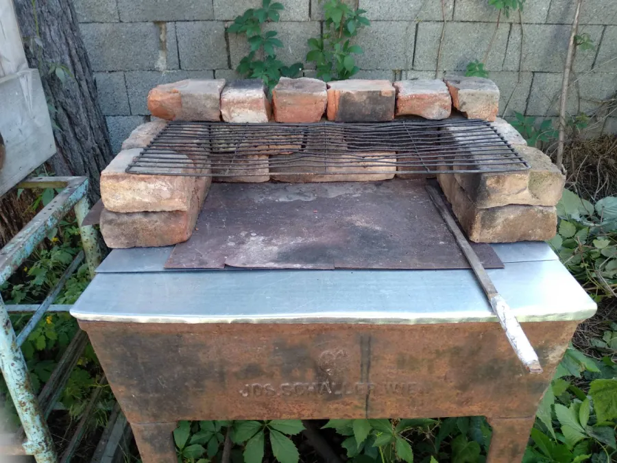 A brick outdoor grill with a metal grid on top, placed in a shady spot against the wall. The grill is on a metal construction with a sheet metal and around it grow green plants.
