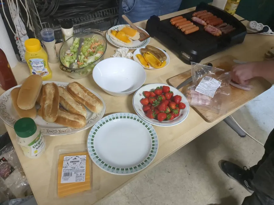On the table is prepared food for informal dining. There are bread, salad, strawberries and cheese slices. On the table are also bottles with ketchup and mustard. The table is also equipped with a grill.