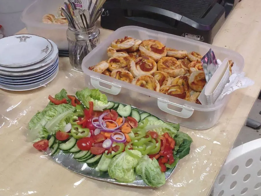 A table with a basket filled with fresh vegetables, including cucumbers, peppers, tomatoes and salad. Next to the table is a box with vegan pizza rolls. In the background, you can see several stacked plates and glasses with utensils.