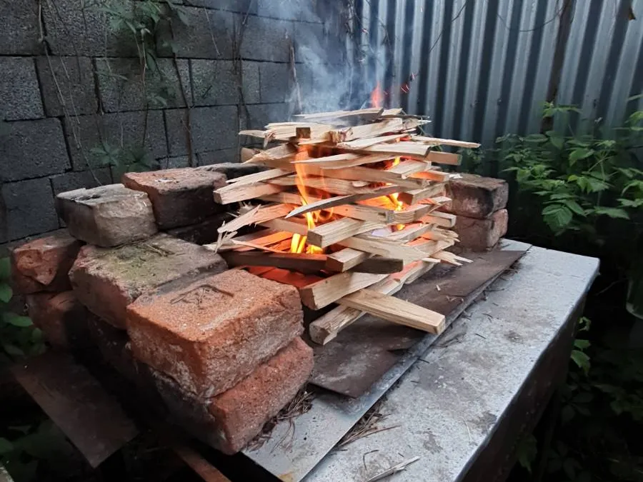 A wooden boundary prepared for lighting on an outdoor grill from brick and metal base. The flames and smoke rise upwards, while in the background you can see a concrete wall and green plants.