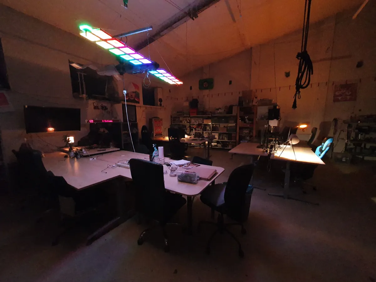Hackerspace Base48 in the evening with a few working tables and computer chairs. The tables are suspended above the tables, serving as LED lights. In the background, you can see the police with equipment and tools.