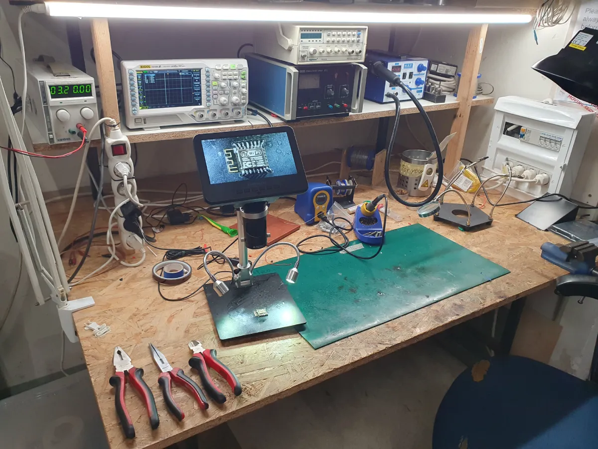 Working table in the hackerspace with various electronic devices, including an oscilloscope, power supply, and soldering iron. The table also has pliers and a microscope with a display showing a detailed view of an ESP32 microchip. The table is well-lit with a fluorescent light above it.