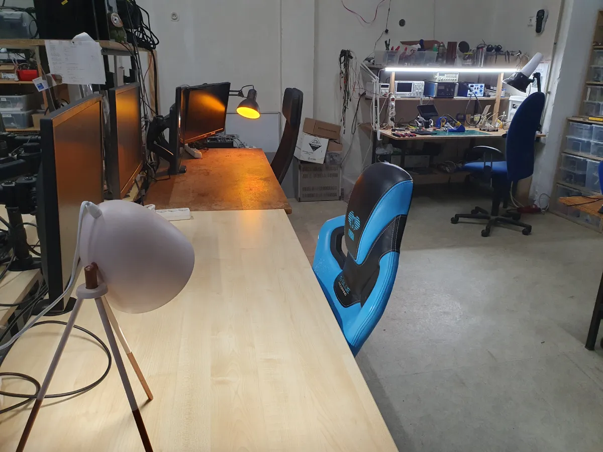 Working space with several tables equipped with computers and monitors, chairs and lamps. In the background, you can see another working table with electronics, tools and measuring instruments.